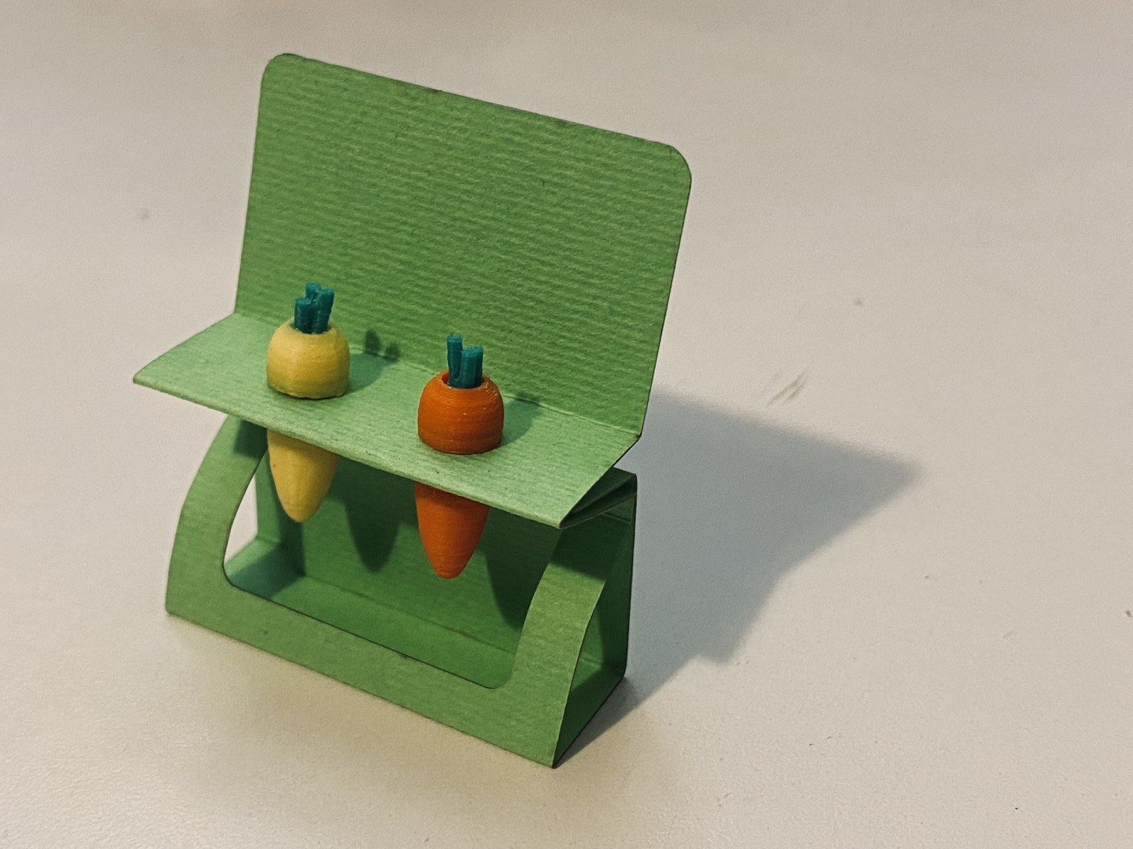 Boucles d'oreilles carottes Holy Sheet, imprimées en 3D : une jaune et une orange à fane turquoise, sur leur support en papier vert découpé